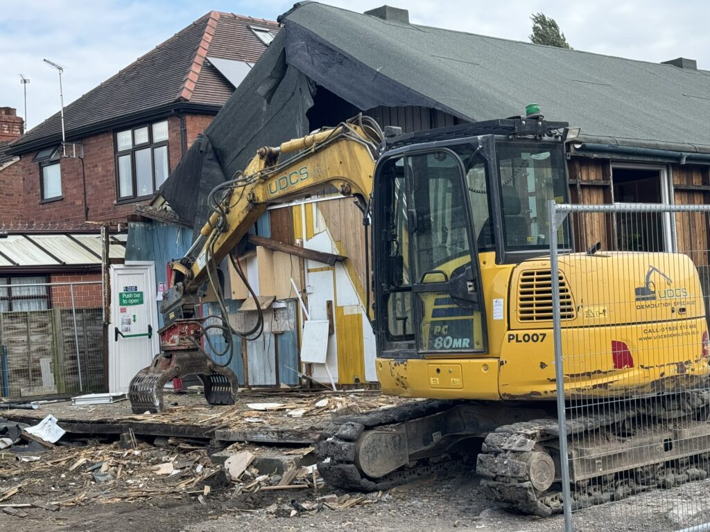 A yellow and black commercial demolition vehicle rests having torn down a section of a wood built Scout hall