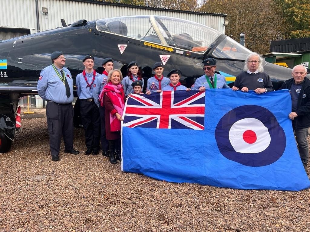 A group of scouts in air scout unfirm stand with a flag of the RAF roundel with the Union flag in the top corner. They stand in front of a fighter plane.