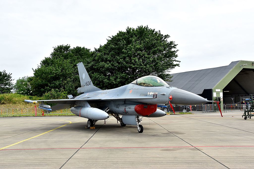 Falcon F-16 aircraft on an air base with trees and a hangar behind.