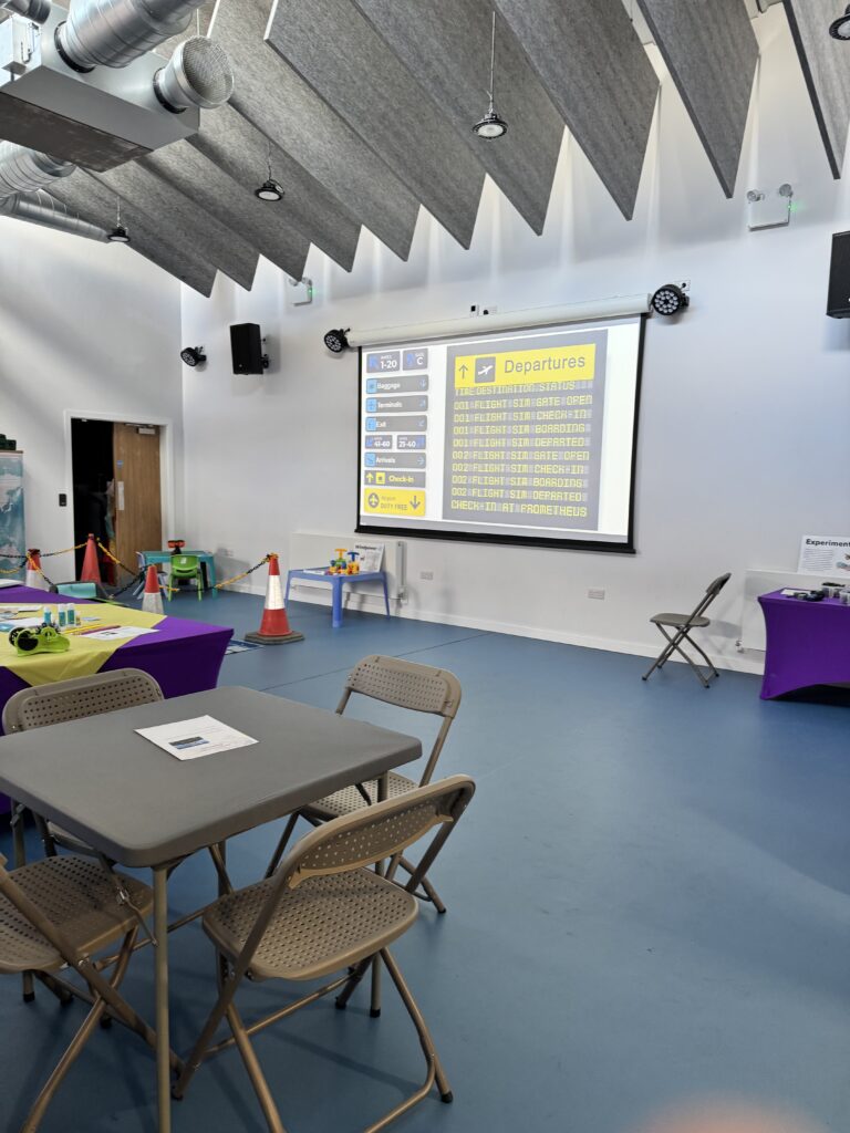 A large meeting hall with a several tables and chairs. A projector screen is shown on one wall. Sound proofing baffle boards hang from the ceiling.