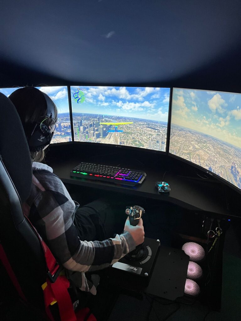 Three screens displaying an aerial view and a young woman controlling a digital plane using foot pedals and joy sticks.